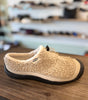A beige sherpa fleece slip-on shoe with a black textured sole, resting on a wooden table with a blurred store background.