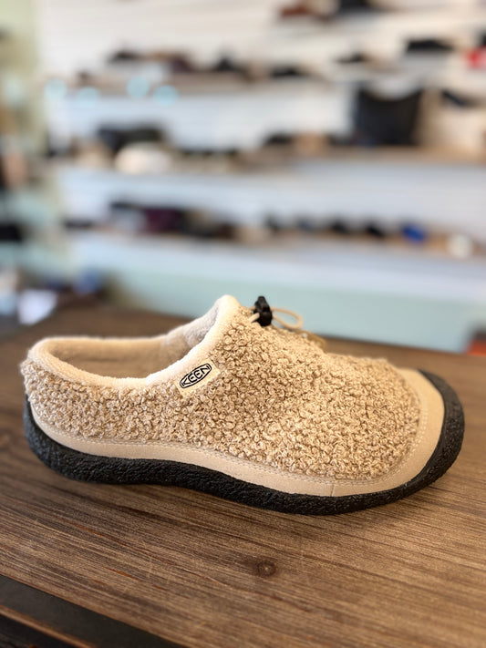 A beige sherpa fleece slip-on shoe with a black textured sole, resting on a wooden table with a blurred store background.